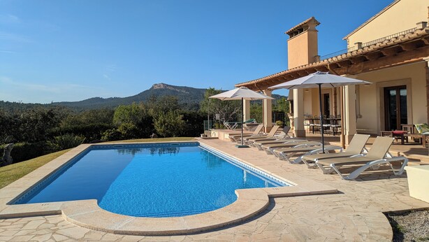 Piscina com espreguiçadeiras e guarda-sóis, montanha ao fundo e um edifício com telhado de cerâmica.