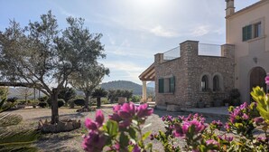 Um edifício de pedra com varanda, rodeado por um jardim com flores roxas e uma árvore.