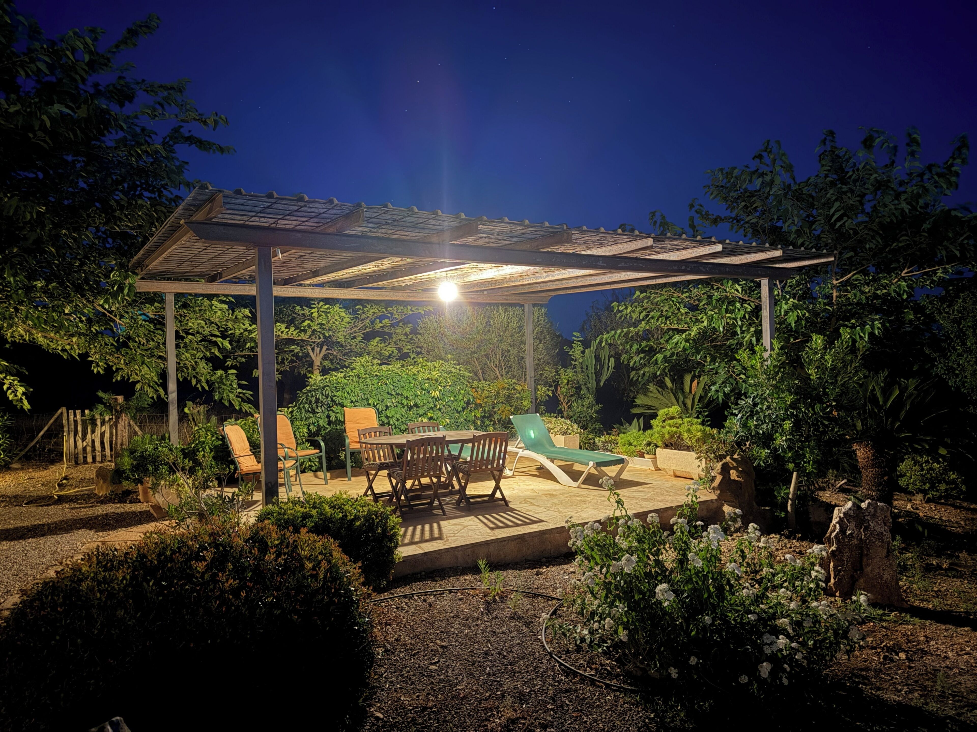 A covered outdoor patio with a table and chairs, surrounded by greenery and a swimming pool.