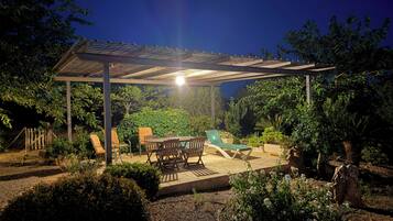 A covered outdoor patio with a table and chairs, surrounded by greenery and a swimming pool.