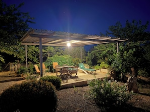 A covered outdoor patio with a table and chairs, surrounded by greenery and a swimming pool.