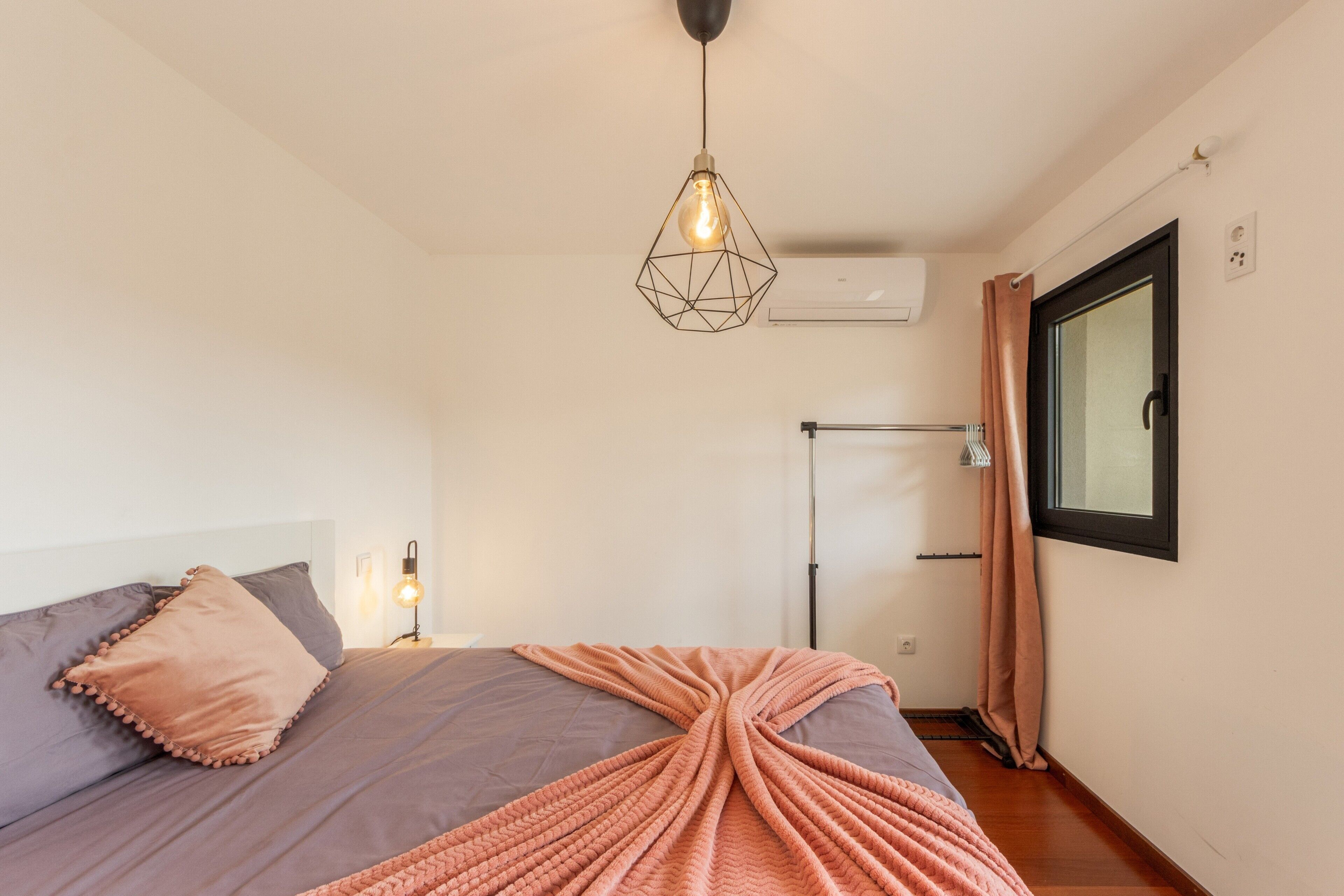 A bedroom with a bed, a hanging light fixture, an air conditioning unit, a window with curtains, and a wall-mounted light.
