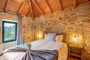 Ein Schlafzimmer mit einer Steinsäule, Holzbalken an der Decke, einem Bett mit Überwurfdecke, zwei Nachttischlampen und einem Fenster mit Vorhängen.
