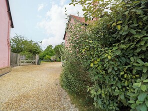 A gravel driveway leading to a pink building with a wooden gate, surrounded by dense green foliage. - The Mallows (Walsingham)