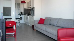 Uma sala de estar moderna com um sofá cinza e almofadas vermelhas, uma poltrona vermelha e uma área de cozinha com armários brancos e uma luminária pendente vermelha.