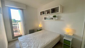 Quarto com cama, janela, radiador, estante com livros e uma pequena mesa com abajur.