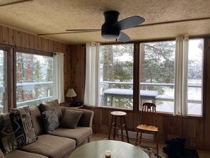 A cozy room with a wooden interior, a ceiling fan, a sofa with cushions, a small table, and a view of a snowy landscape through large windows. - Baysong Cottage, with Sauna, on Georgian Bay. (MacTier)