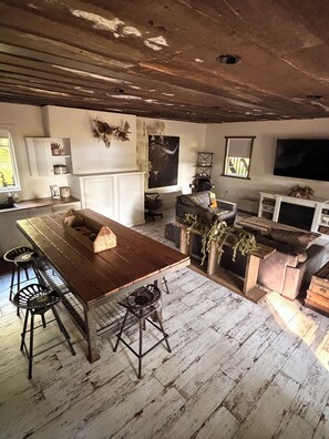 A rustic interior with a wooden dining table, bar stools, a sofa, and a television.