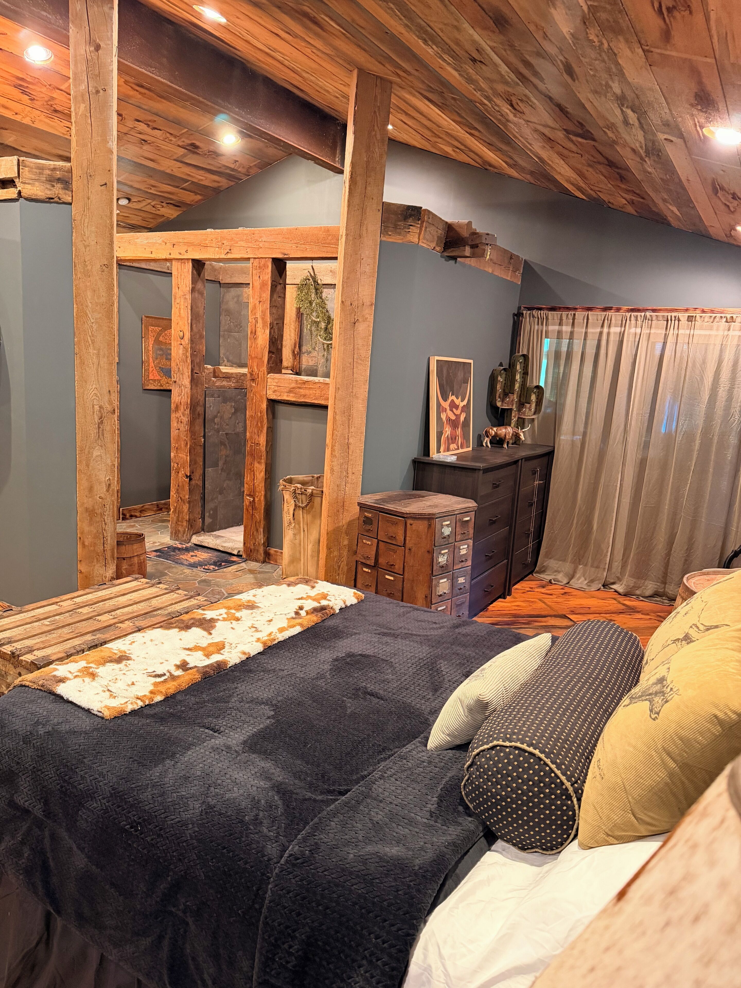 A room with a bed, wooden beams, a stone fireplace, and a window with curtains.