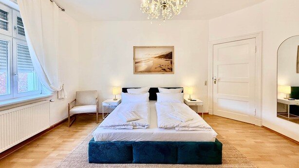 A bedroom with a large bed, two bedside tables with lamps, a chandelier, a painting on the wall, and a window with curtains. - Beautiful 3-bedroom Apartment 'Johann' (Stendal)