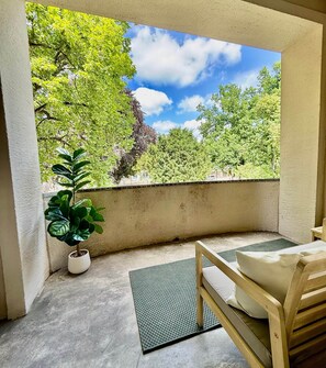 Ein Balkon mit Blick auf Bäume und Himmel, eine Topfpflanze, eine Fußmatte und ein Holzstuhl.