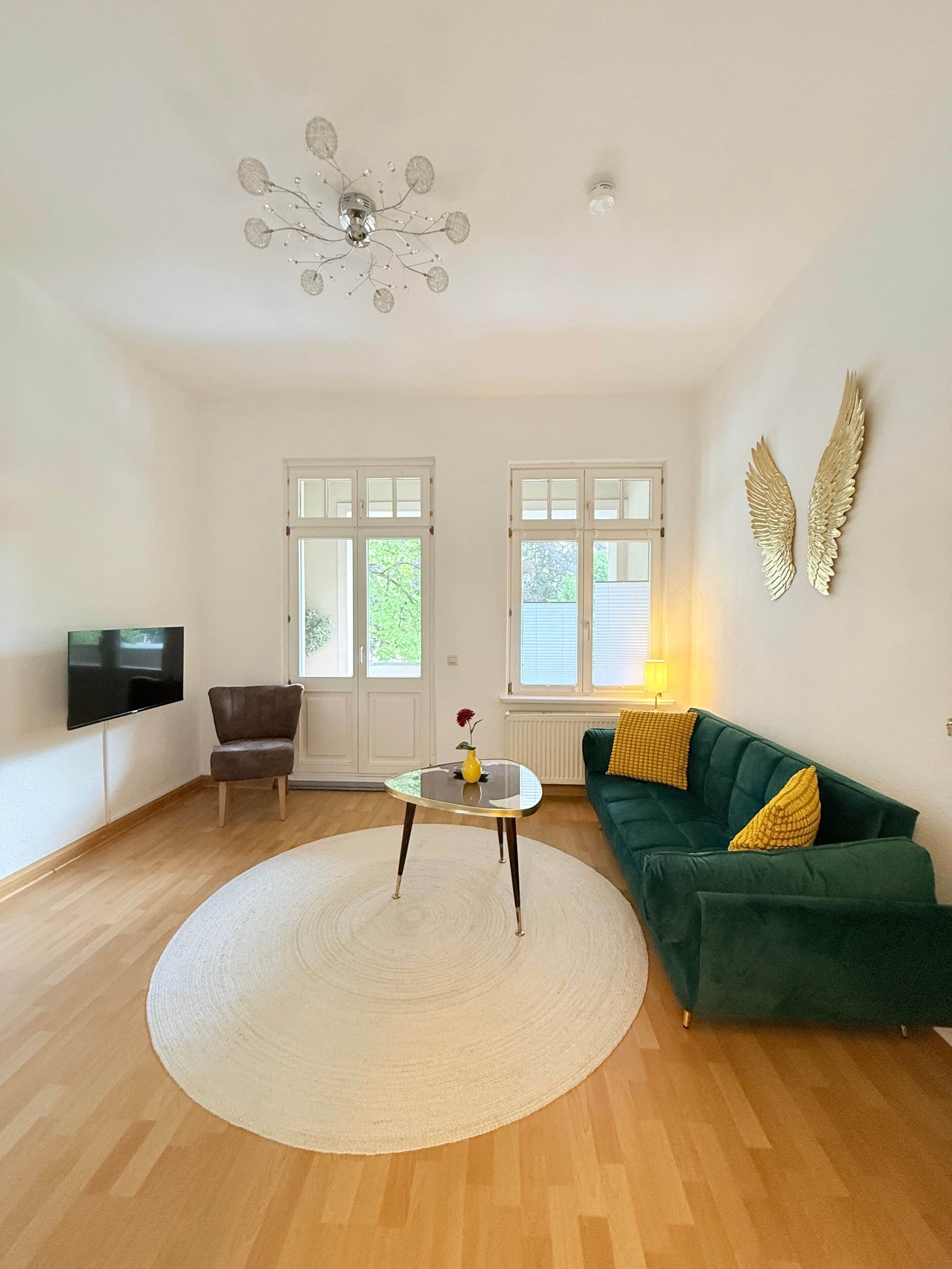 Ein großzügiges Wohnzimmer mit einem grünen Sofa, einem runden Couchtisch, einem an der Wand montierten Fernseher und einer dekorativen Deckenleuchte.