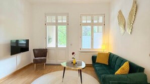 A spacious living room with a green sofa, a round coffee table, a TV mounted on the wall, and a decorative ceiling light. - Beautiful 3-bedroom Apartment 'Johann' (Stendal)