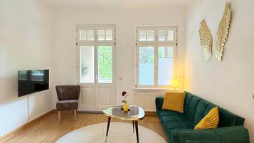 Ein großzügiges Wohnzimmer mit einem grünen Sofa, einem runden Couchtisch, einem an der Wand montierten Fernseher und einer dekorativen Deckenleuchte.