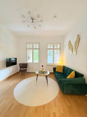 Ein großzügiges Wohnzimmer mit einem grünen Sofa, einem runden Couchtisch, einem an der Wand montierten Fernseher und einer dekorativen Deckenleuchte.