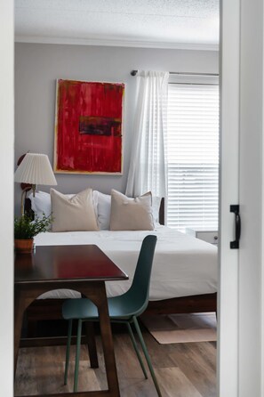 Ein Schlafzimmer mit Bett, Schreibtisch, Stuhl, Lampe, einem roten abstrakten Gemälde und einem Fenster mit Jalousien.