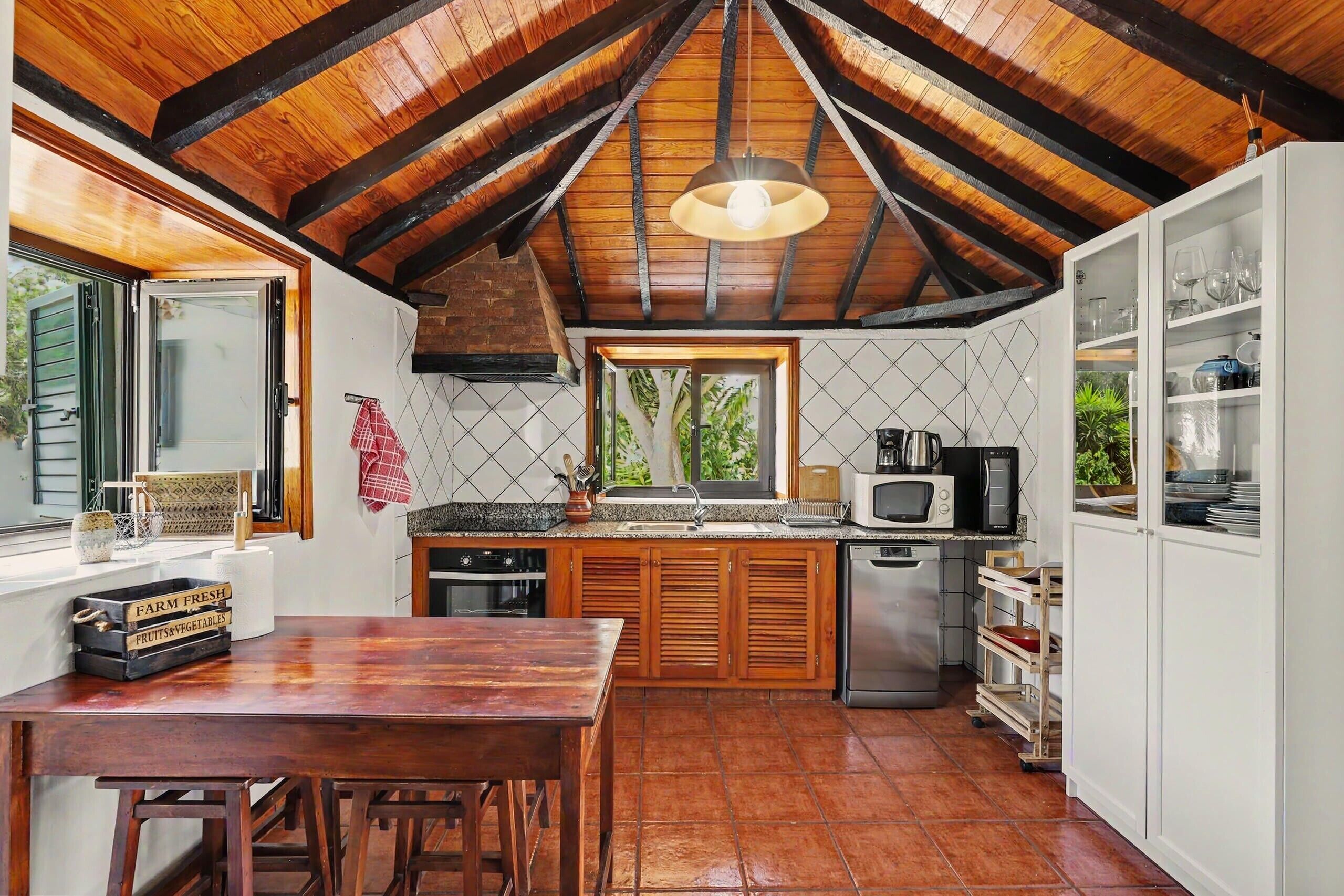 A rustic kitchen with wooden beams, a farmhouse-style table, and a view of greenery outside.