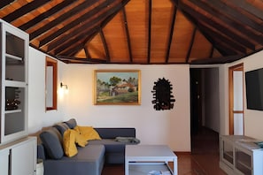 Uma sala de estar com teto de madeira, um sofá cinza com almofadas amarelas, uma mesa branca e um quadro na parede.