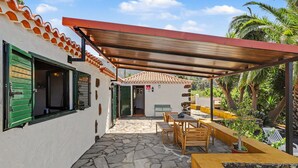 A covered patio with stone flooring, wooden furniture, and a view of a white building with green shutters. - Apartment 'Tocando El Cielo' with Mountain View, Private Terrace and Wi-Fi (Barrial de Arriba)