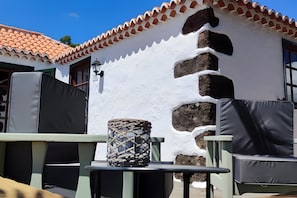 Pátio com mobiliário externo, um enfeite de mesa tecido e um edifício branco com telhado ladrilhado.