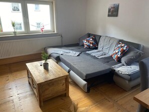 Un espacio habitable compacto con un sofá, una mesa de centro de madera y una ventana con vistas a edificios.