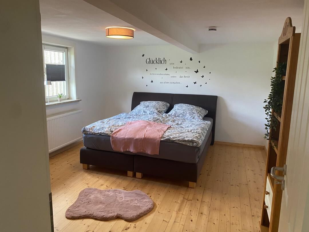 Ein Schlafzimmer mit einem Bett, einer Wandleuchte, einem Fenster mit Jalousien und einer kleinen Pflanze auf einem Regal.