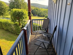 Eine Holzterrasse mit Geländer, zwei Stühlen und Blick auf eine grüne Landschaft mit Bäumen und Hügeln.