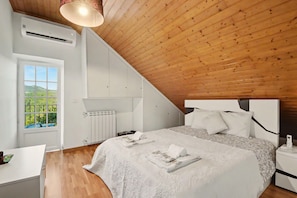 Ein Schlafzimmer mit schrägen Wänden, einem Bett mit weißen Bettwäsche, einem Nachttisch und Blick auf Grünfläche draußen.