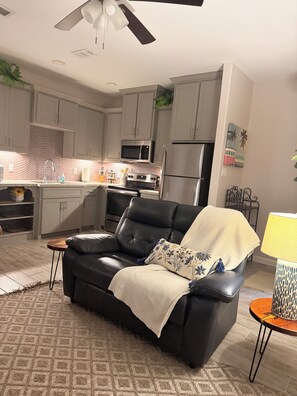 Ein Wohnzimmer mit einer schwarzen Ledersofa, einem gemusterten Teppich, einem kleinen Holztischchen, einer Lampe und einer Küche im Hintergrund.