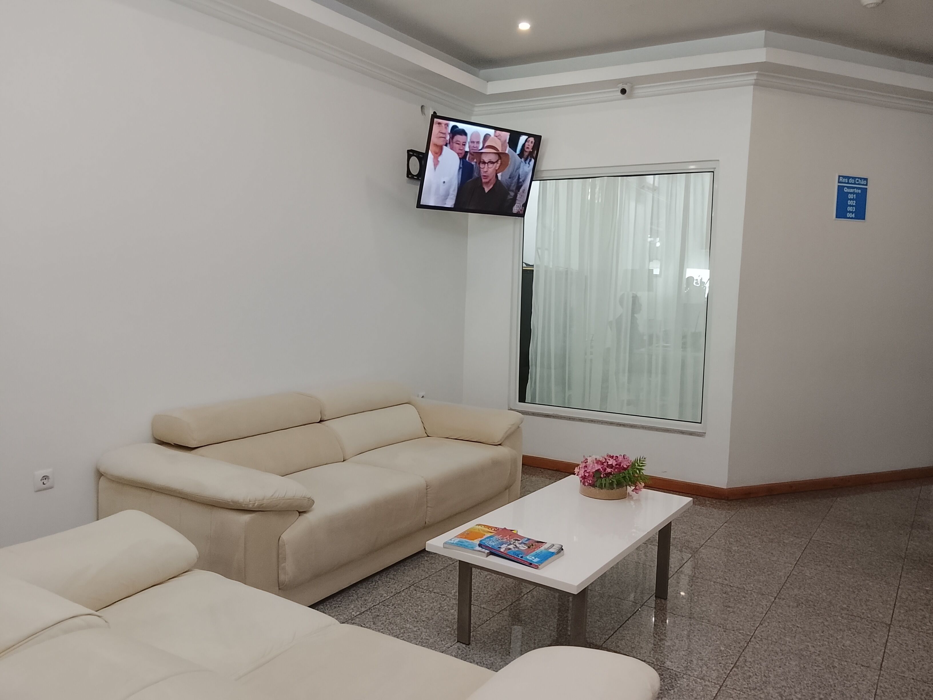 Un área de espera con un sofá, una mesa baja con un libro, un televisor montado en la pared y una ventana con cortinas.