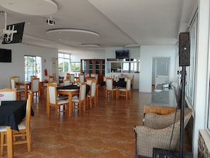 Un grand espace Ă manger avec des tables et des chaises en bois, un coin cuisine dotĂ© dâun comptoir et dâun Ă©vier, et un tĂ©lĂ©viseur fixĂ© au mur.