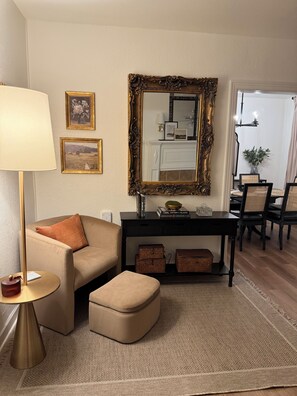 Ein Wohnzimmer mit Sofa, Hocker, Lampe, Wandspiegel, gerahmten Bildern und Kamin.