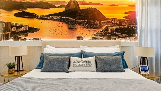Habitación de hotel con cama, almohadas, mesita de noche, lámpara y un cuadro de un paisaje urbano en la pared.