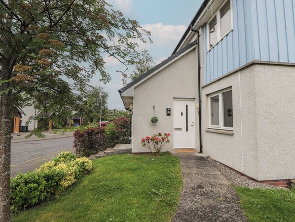 A two-story building with a white exterior, a blue window, and a small garden with flowers. - 39 Knocknagael (Inverness)