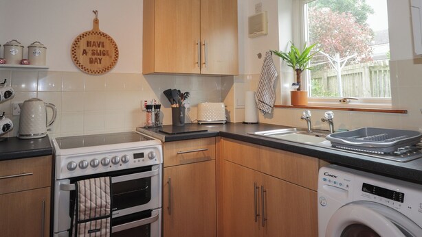 A kitchen with wooden cabinets, a stove, a washing machine, a sink, and a window with a plant. - 39 Knocknagael (Inverness)