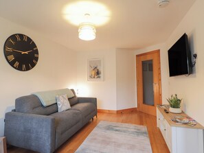 A living room with a sofa, a wall clock, a ceiling light, a framed picture, a door, and a small table with a plant. - 39 Knocknagael (Inverness)