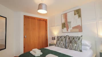 Een net opgemaakt bed met een groene sprei, een houten deur, een aan de muur hangend abstract schilderij en een bedlampje.
