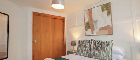 Una cama bien hecha con una colcha verde, una puerta de madera, un cuadro abstracto colgado en la pared y una lámpara de noche.