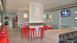 A modern bar area with red stools, a wooden counter, and a TV screen displaying a news program. - Eklo Hotels Le Mans (Le Mans)