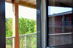 A window with a metal grid, a wooden beam, and a view of green foliage and a building.