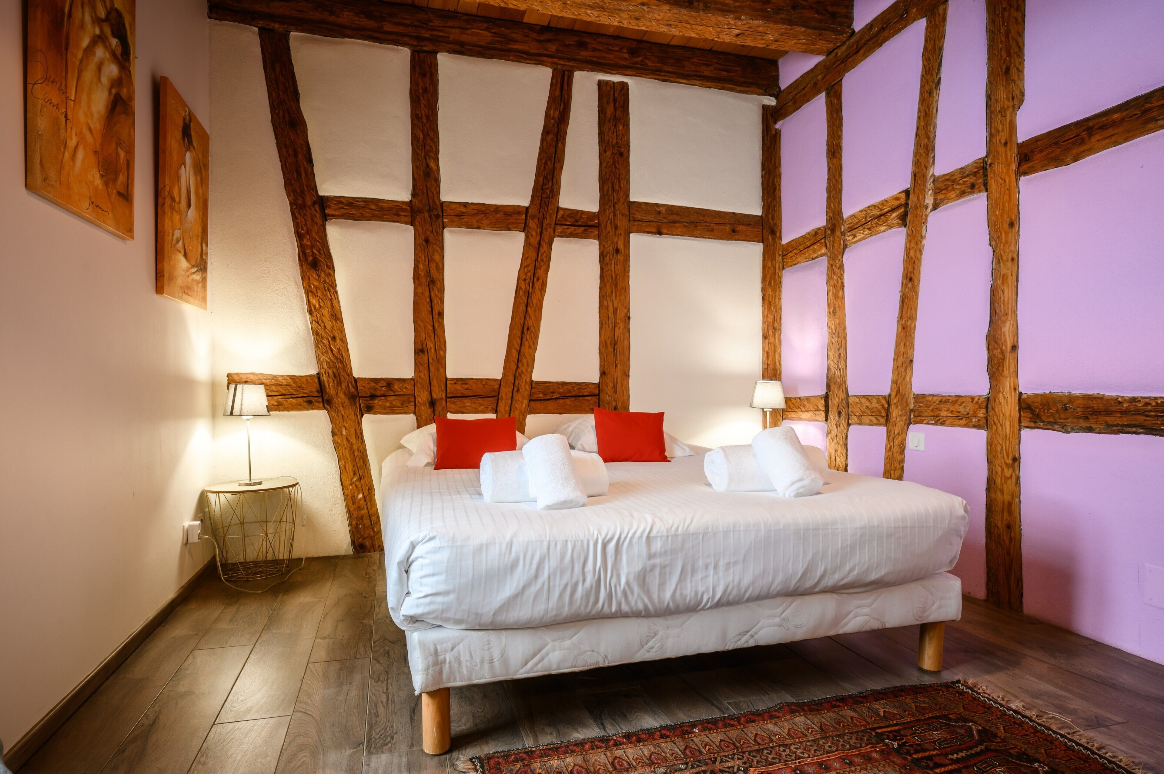 Een slaapkamer met een groot bed, houten balken, een klein nachtkastje en een vloerkleed met dessin.
