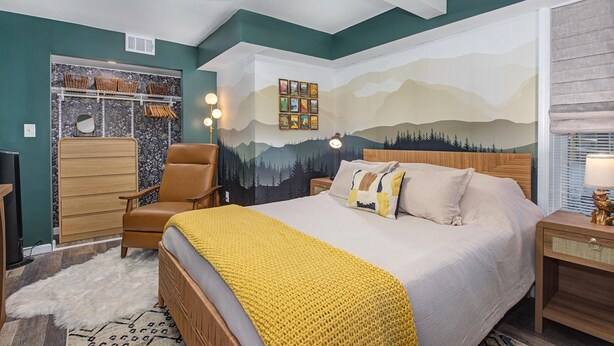 Un dormitorio con cama, una silla, un tocador, un televisor y una pared decorada con un mural de montañas.