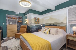 Ein Schlafzimmer mit Bett, Sessel, Kommode, Fernseher und einer Wand mit Bergbild.