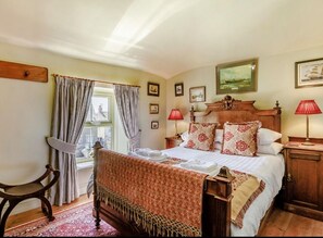A traditional bedroom with a bed, bedside lamps, a chair, a window with curtains, and a view of buildings outside.