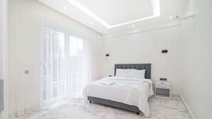 Un dormitorio con cama, mesitas de noche, una lámpara en el techo y un amplio ventanal con cortinas.