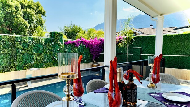 Mesa de vidrio dispuesta con servilletas rojas, copas de vino y botellas, con vista a una piscina y exuberante follaje.