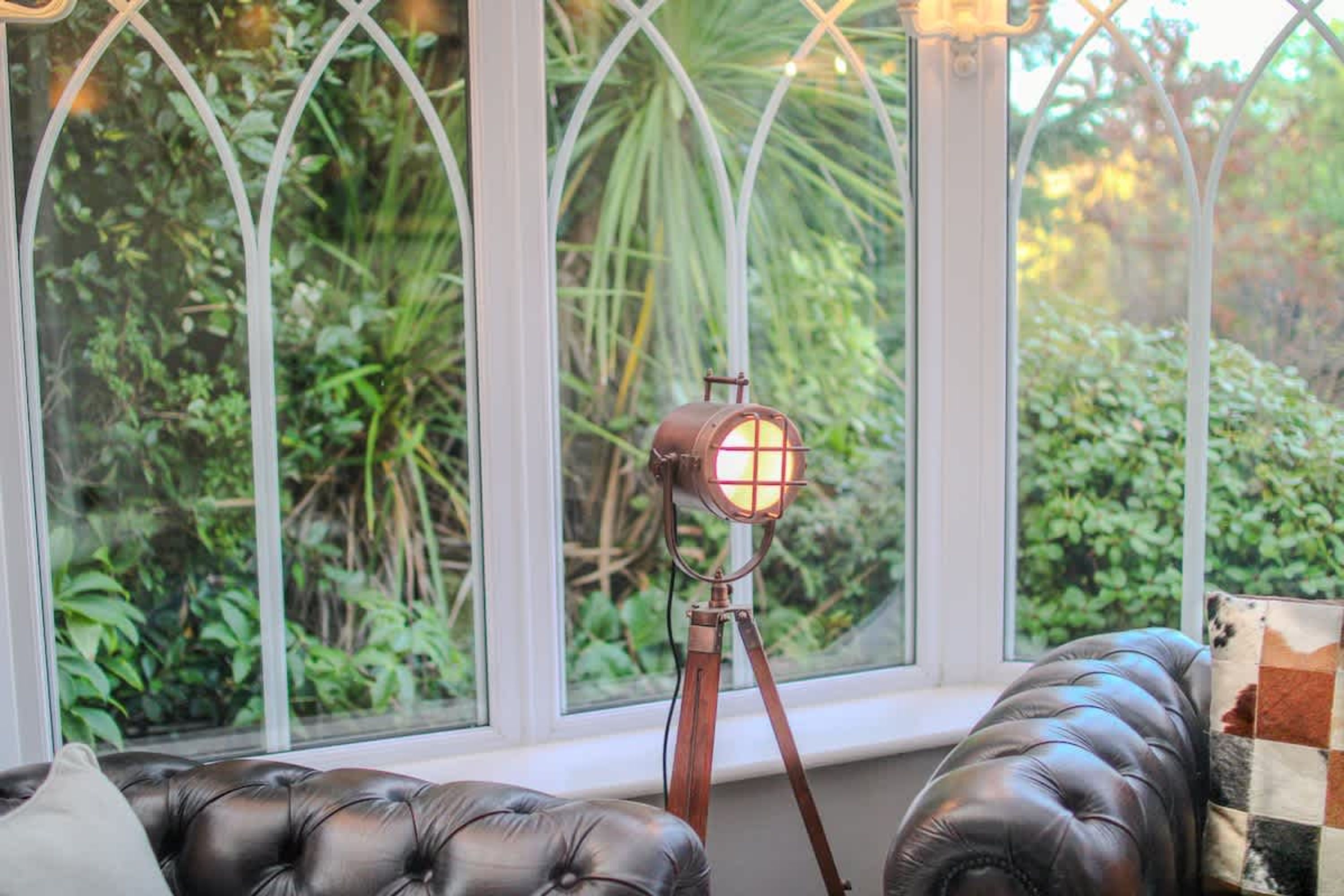 A room with a leather sofa, a tripod lamp, and a view of lush greenery through large windows.