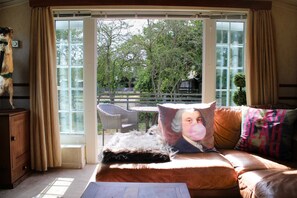 A room with a sofa, a window with curtains, a view of the outdoors, and a small table. - Berney Lodge - Puppy Lounge Arcades Games Room (Market Rasen)