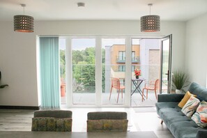 Una sala moderna con un sofá, otomanas, una mesa de comedor con sillas, y un balcón con vistas a edificios y zonas verdes.