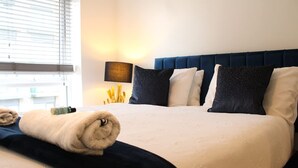 Una cama bien hecha con cabecera azul, una lámpara de noche y una ventana con persianas.